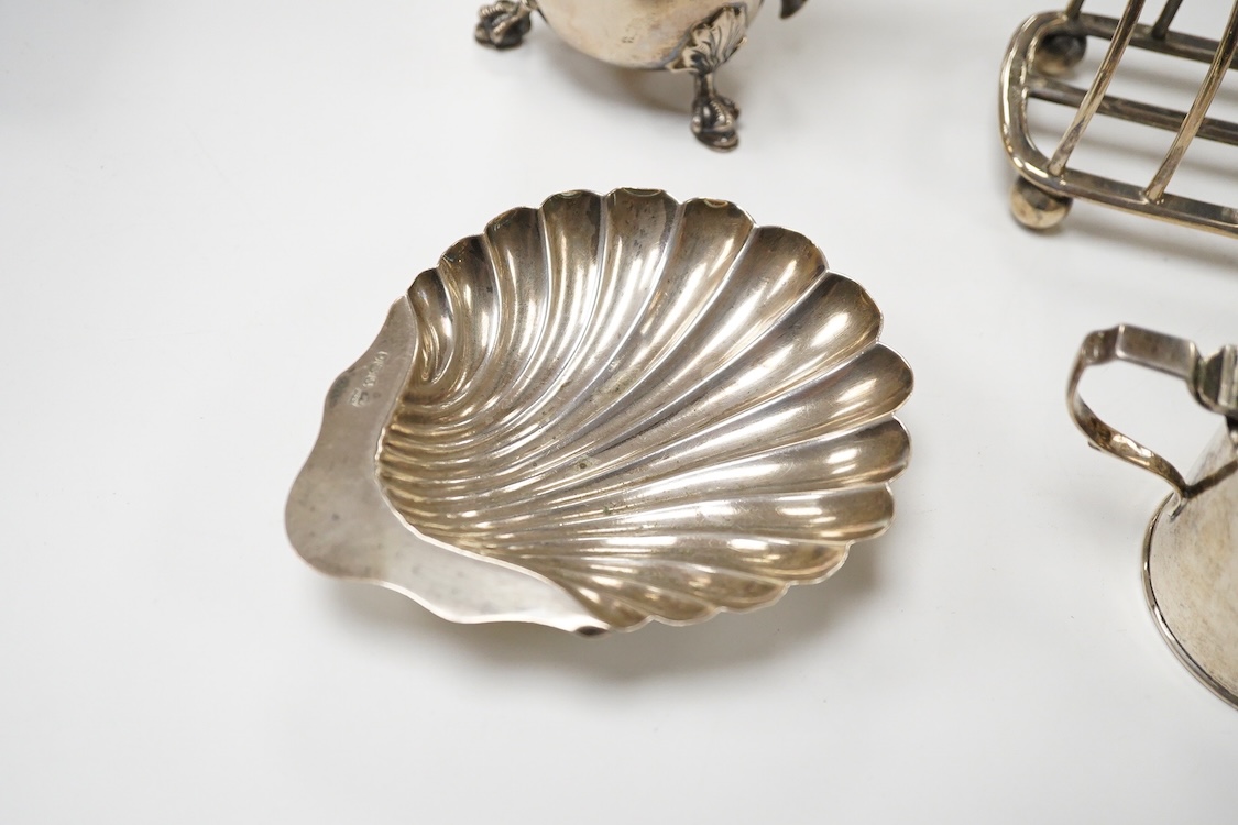 An Edwardian silver mug, on claw and hoof feet, William Hutton & Sons, London 1909, height 83mm, together with a silver butter shell, mustard pot and a silver five bar toastrack, 17oz.